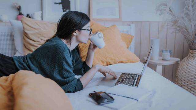 A foto mostra uma mulher deitada na cama, tomando café e mexendo no notebook, pesquisando sobre book de ofertas