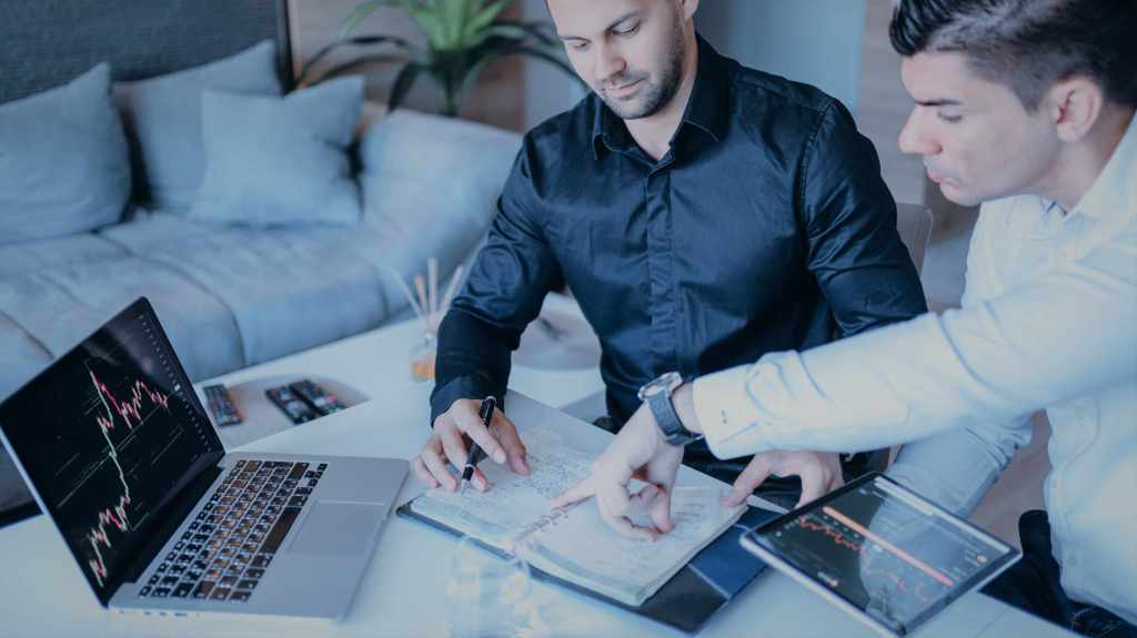 Dois homens sentados em frente a uma mesa, observando anotações em um caderno. Um deles segura um tablet exibindo gráficos de ações. À frente dos dois há um notebook exibindo outro gráfico.