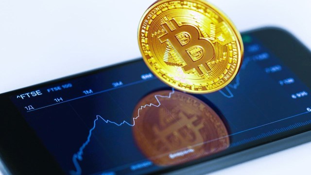 Uma moeda que representa o Bitcoin apoiada sobre um celular que o rendimento de um ETF de criptomoedas ao longo do dia.