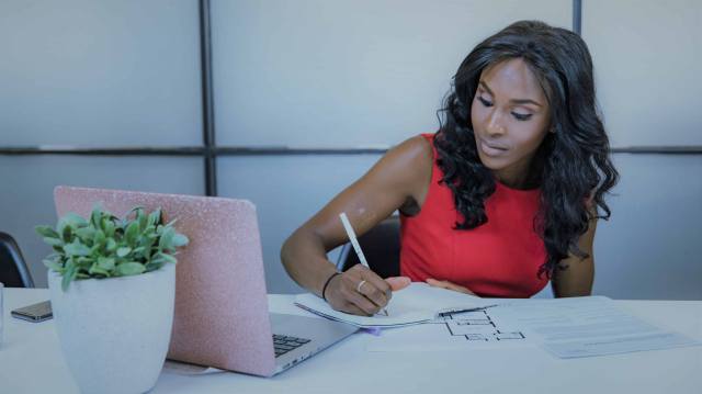 A foto mostra uma mulher, em um escritório, fazendo anotações sobre média móvel simples em um caderno. À sua frente, na mesa, ainda há um notebook e um vaso de plantas.