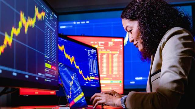 Mulher usa diversos monitores que exibem gráficos de ações, analisando as tendências das ações da Petrobras.