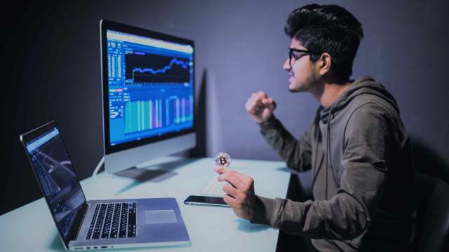 A imagem mostra um homem sentado de frente para um notebook e uma tela de computador que mostra um gráfico do mercado financeiro em referência ao momento do pregão na bolsa.