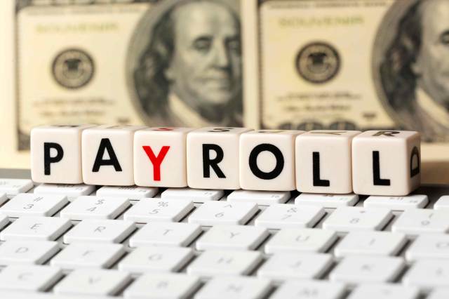 Imagem mostra um teclado e dados escritos "payroll" e uma nota de dólar americano ao fundo.