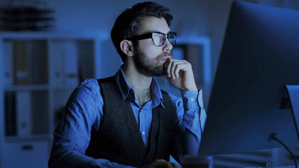 A imagem mostra um homem de barba sentado de frente para a tela de um computador com a mão no queixo, com tom de pensativo, como se estivesse identificando um candle de força. Ele está usando um óculos preto e camisa social azul com um colete preto por cima.