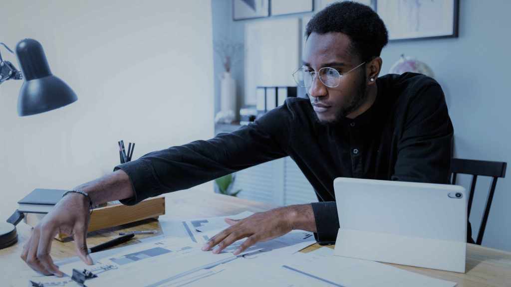 A imagem mostra um homem negro de camisa preta e óculos sentado em uma mesa de escritório. Ele está pegando alguns papéis da mesa e em cima também há um tablet, canetas e cadernos, em referência ao estudo e cálculo do grau de alavancagem operacional.