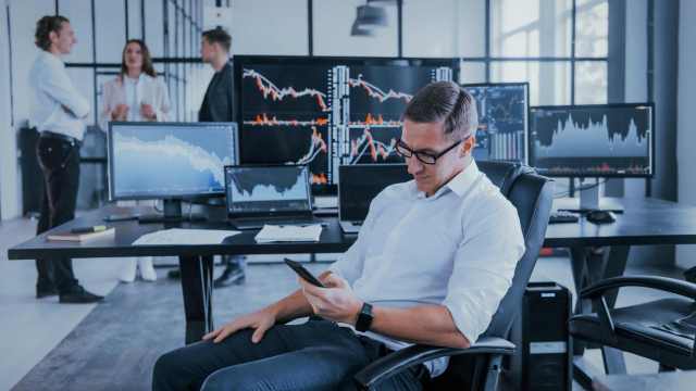 A imagem mostra um homem branco de camisa branca, calça preta e óculos. Ele está sentado em uma cadeira olhando para o celular em sua mão. Atrás dele temos uma mesa com notebooks e monitores que mostram vários gráficos de ações, em referência a análise do engolfo de alta.