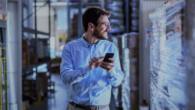 A imagem mostra um homem andando de frente segurando um celular olhando para o lado sorrindo. Ele veste uma camisa social, está de óculos e o seu dedo aponta para a tela do celular em referência ao entendimento sobre o indicador estocástico.