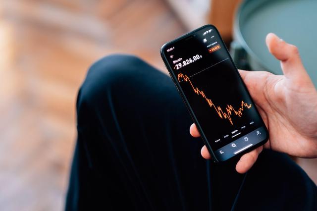 Imagem com foco em uma pessoa segurando um celular acompanhando um gráfico em um aplicativo de trading.
