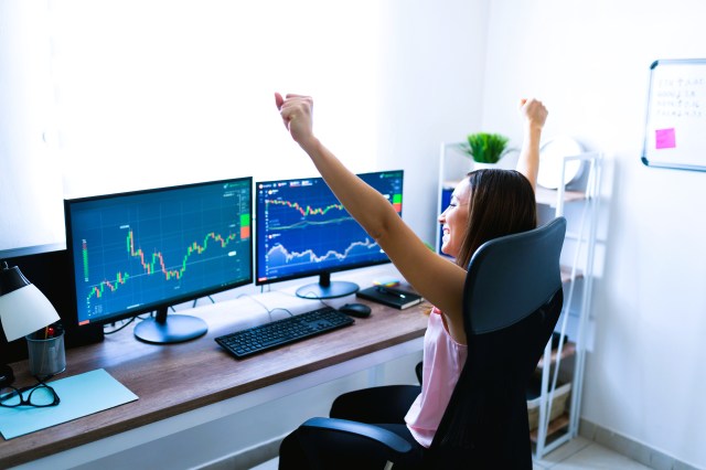 Mulher sentada à frente de dois monitores com gráficos de ações, comemorando com os braços para cima.