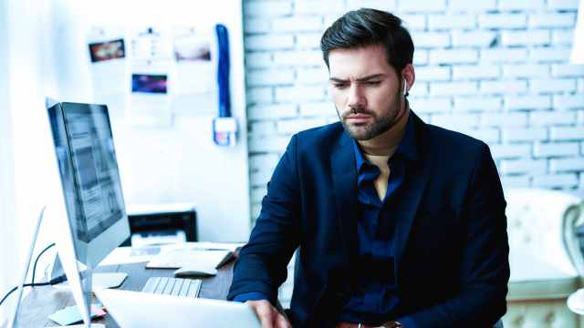 A imagem mostra um homem de barba, com o cabelo curto penteado para o lado, vestindo uma camisa social e terno azul marinho. Ele está em uma mesa de escritório, sentado usando um notebook com uma cara séria e de preocupação em referência ao viés de confirmação no trading.