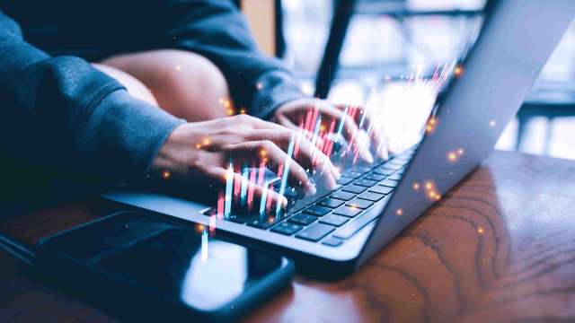 A imagem mostra as mãos de uma pessoa mexendo no notebook em cima de uma mesa ao lado de um celular. Também podemos ver gráficos de candles saindo da tela em relação ao tema de reversão de tendência e day trading.