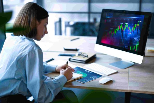 Pessoa sentada à mesa com computador mostrando gráficos financeiros, fazendo anotações para entender como investir na bolsa de valores.