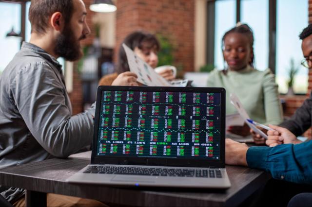 Notebook em cima de uma mesa com códigos de ações à mostra. Atrás, um grupo de pessoas discute sobre os significados e os tipos de ações.