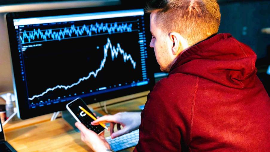 Homem usando capuz vermelho e segurando um celular analisa gráfico de ações com indicador MACD na tela do computador.