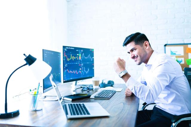 Homem trader em frente a telas com gráficos do mercado de ações, comemorando um resultado positivo após o Trade Assistido.