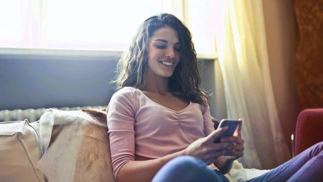 Mulher sentada num sofá, com expressão sorridente, pesquisando em seu celular como a taxa referencial pode afetar seus investimentos.
