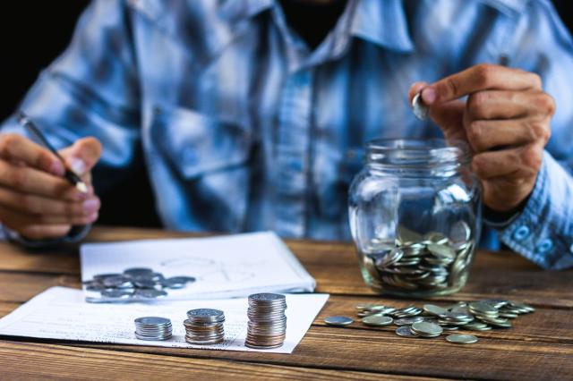 Pessoa sentada em frente a uma mesa, com lápis, papéis e moedas empilhadas, decidindo onde investir o décimo terceiro salário.