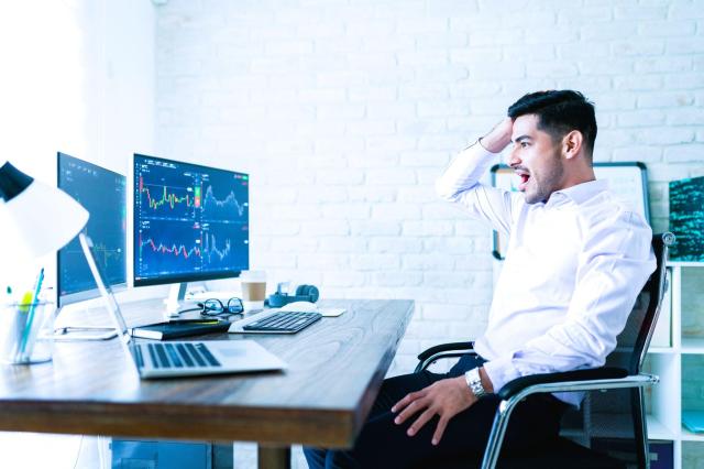Homem surpreso, em frente ao computador com gráficos da bolsa, para exemplificar a importância da psicologia do trader.