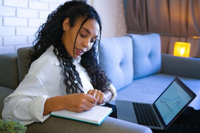Mulher sentada no sofá, com um notebook no colo para pesquisar sobre a restituição do IR no day trade, enquanto faz anotações em um caderno.