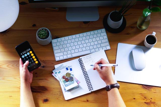 Mesa vista de cima, com computador, cadernos e notas de dinheiro. É possível ver os braços de uma pessoa, segurando um celular com a calculadora aberta e anotando contas relacionadas à restituição do day trade.