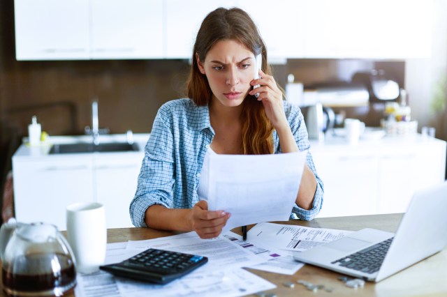Mulher branca com uma expressão de preocupação, segurando um celular na orelha, sentada em frente à uma mesa com papéis espalhados, calculadora e um notebook. 