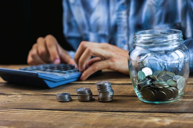 Pessoa utilizando calculadora para simular rendimentos em renda fixa e variável com pilhas de moedas e pote de economia sobre a mesa.
